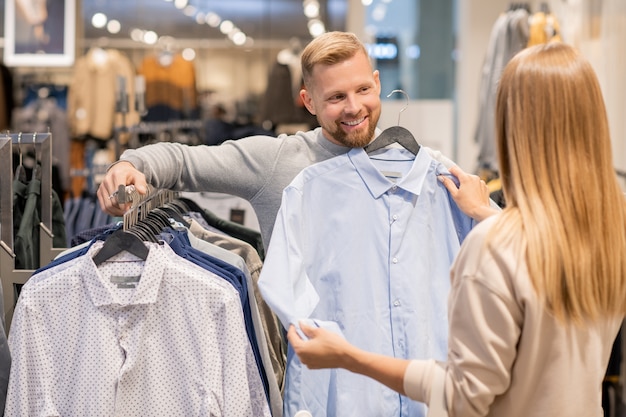 Casal jovem escolhendo uma nova camisa para homem enquanto fica em pé no rack com a nova coleção no departamento de roupas do shopping