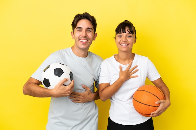 Casal jovem esportivo jogando futebol e basquete isolado em fundo amarelo sorrindo muito enquanto coloca as mãos no peito