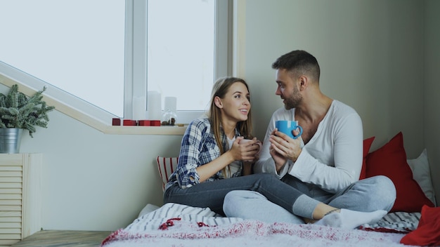 Casal jovem feliz conversando e rindo enquanto está sentado na cama e bebe café pela manhã