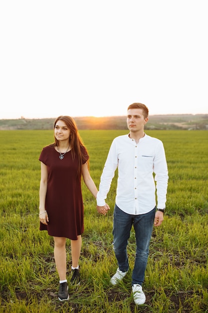 Casal jovem, feliz e amoroso, ao pôr do sol, de pé em um campo verde, contra o céu, de mãos dadas e curtindo um ao outro