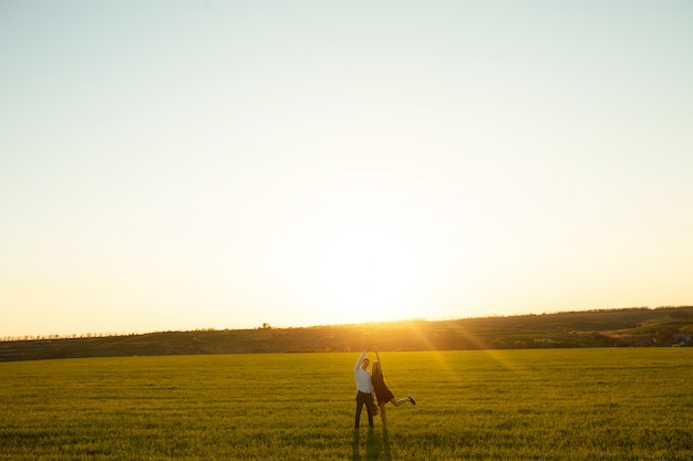 Casal jovem, feliz e amoroso, ao pôr do sol, de pé em um campo verde, contra o céu, de mãos dadas e curtindo um ao outro