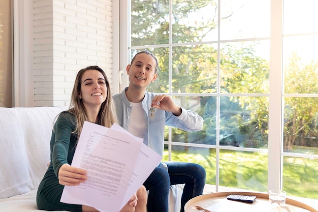 Casal jovem feliz e sorridente mostrando o contrato de assinatura e as chaves de sua nova casa sentada em t