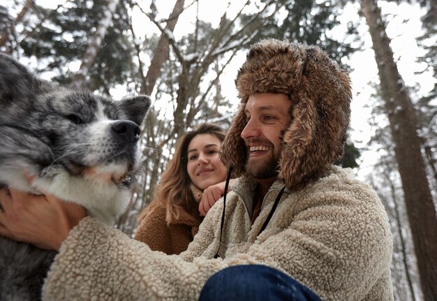 Casal jovem feliz em roupas de inverno brincando com husky siberiano de raça pura e se divertindo na floresta de pinheiros de neve