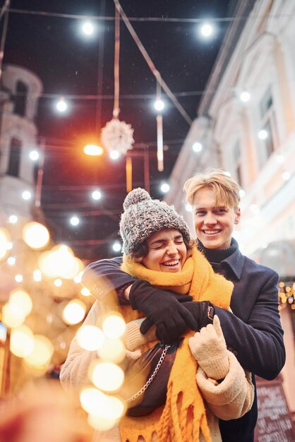 Casal jovem feliz em roupas quentes está juntos na rua decorada de Natal.