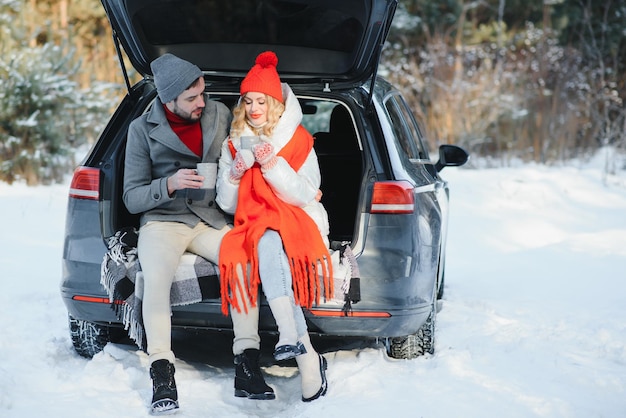 Casal jovem feliz está sentado no porta-malas do carro na floresta de inverno e bebendo chá quente da garrafa térmica. inverno