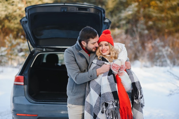 Casal jovem feliz está sentado no porta-malas do carro na floresta de inverno e bebendo chá quente da garrafa térmica. inverno