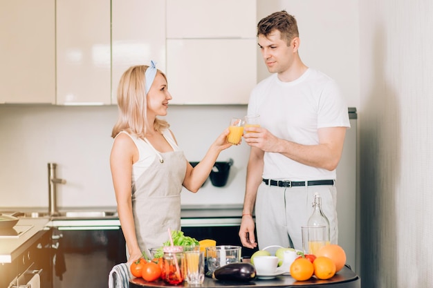 Casal jovem feliz fazendo bebida orgânica na cozinha juntos, está batendo os copos, bebendo suco de laranja espremido na hora na cozinha, comemorando o sucesso do marido no trabalho. Conceito de estilo de vida saudável.