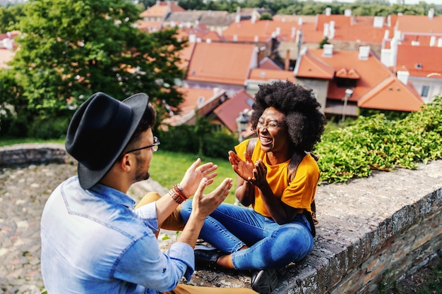 Casal jovem feliz hippie sentado ao ar livre em uma parte velha da cidade, sorrindo e flertando.