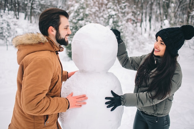 Casal jovem feliz no inverno. Família ao ar livre. homem e mulher olhando para cima e rindo. Amor, diversão, estação e pessoas - caminhando em winter park. Fazendo um boneco de neve.