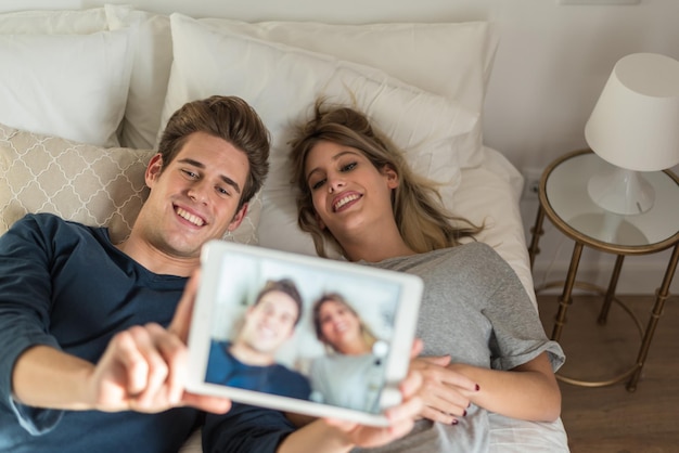 Casal jovem feliz relaxou na cama, tirando uma selfie com o tablet