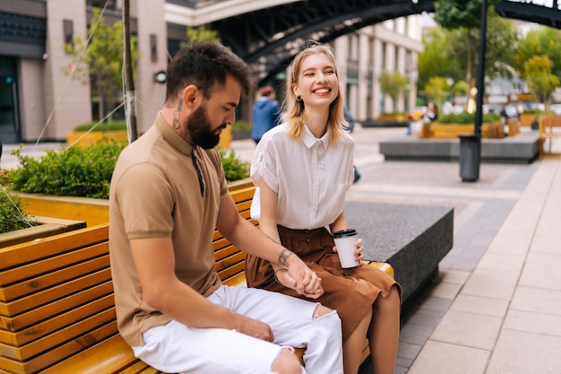 Casal jovem feliz sentado no banco e bebendo café para viagem aproveitando o tempo juntos ao ar livre no verão