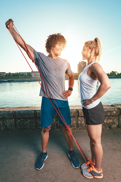 Casal jovem fitness está fazendo exercícios com elástico à beira do rio em um pôr do sol. O homem está esticando os braços e a mulher o apoia.