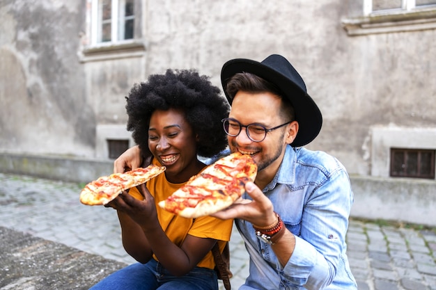 Casal jovem fofo multicultural hippie sentado ao ar livre e comendo pizza.