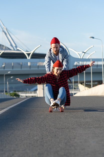 Casal jovem hippie se divertindo em longboard ao ar livre, andando na estrada da cidade, rindo juntos