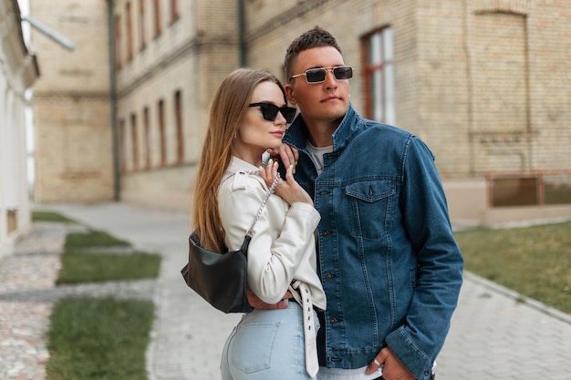 Casal jovem hipster legal na moda em roupas da moda com óculos de sol da moda em roupa e uma bolsa andando na cidade Cara bonito em uma jaqueta jeans e garota elegante com uma jaqueta de couro branca