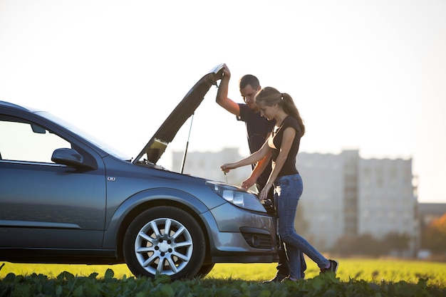 Casal jovem, homem bonito e mulher atraente no carro com capô estourado, verificando o nível de óleo no motor usando a vareta no céu claro. transporte, problemas de veículos e conceito de avarias.