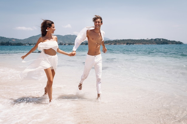 Casal jovem, homem e mulher bonita em roupas brancas, correndo ao longo da praia