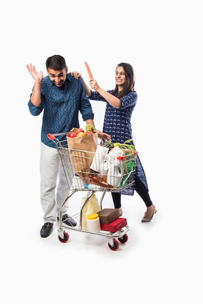 Casal jovem indiano com carrinho de compras ou carrinho cheio de mantimentos, legumes e frutas. foto de corpo inteiro isolada sobre uma parede branca