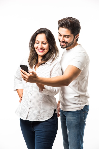 Foto casal jovem indiano usando smartphone ou aparelho celular, isolado sobre um fundo branco ou de tijolo vermelho