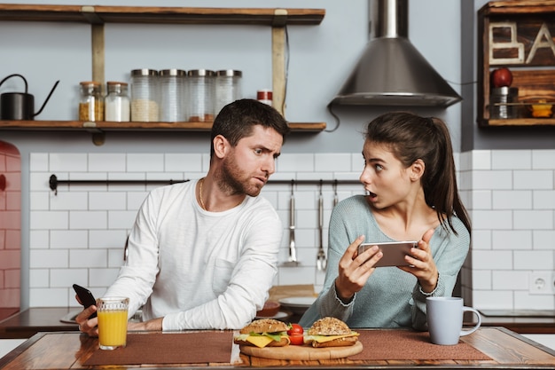 Casal jovem infeliz sentado na cozinha durante o almoço em casa, tendo um problema, segurando um telefone celular