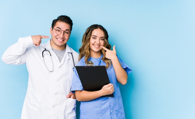 Casal jovem médico posando em uma parede azul isolada sorrisos, apontando os dedos na boca.