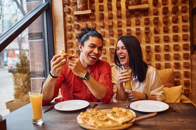 Casal jovem multiétnico sentados juntos dentro de casa e comendo pizza.