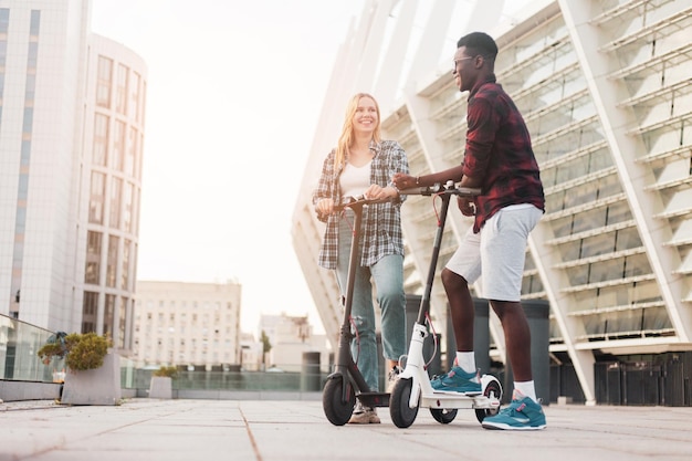Casal jovem multirracial andando de scooters elétricos na cidade e sorrindo