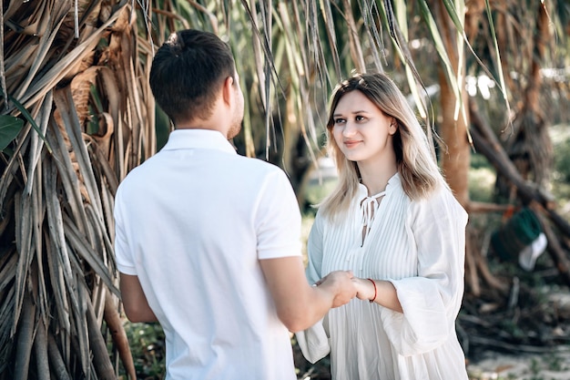 Casal jovem na moda ativo com roupas brancas está em pé na praia e de mãos dadas. Loira linda com fio vermelho no braço e um homem estão de férias. Phuket. Tailândia