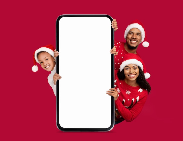 Casal jovem negro feliz e criança pequena em chapéus de Papai Noel em grande smartphone com tela vazia em fundo vermelho