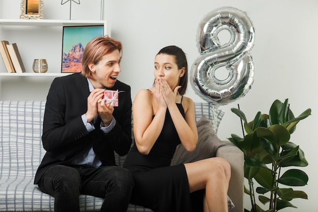 Casal jovem no dia da mulher feliz sorrindo e dando um presente para uma garota surpresa sentada no sofá da sala.