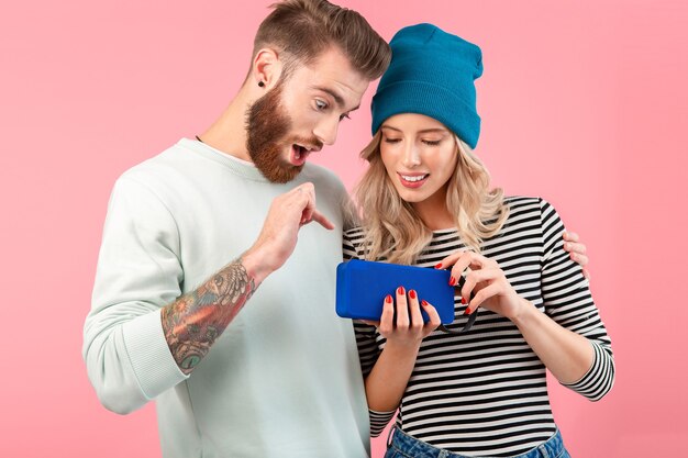 Casal jovem ouvindo música no alto-falante sem fio, vestindo roupas elegantes e legais, sorrindo, posando em rosa