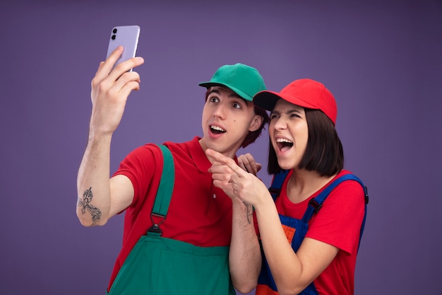 Casal jovem rapaz animado e garota alegre com uniforme de trabalhador da construção civil e boné levantando o celular, olhando e apontando para ele