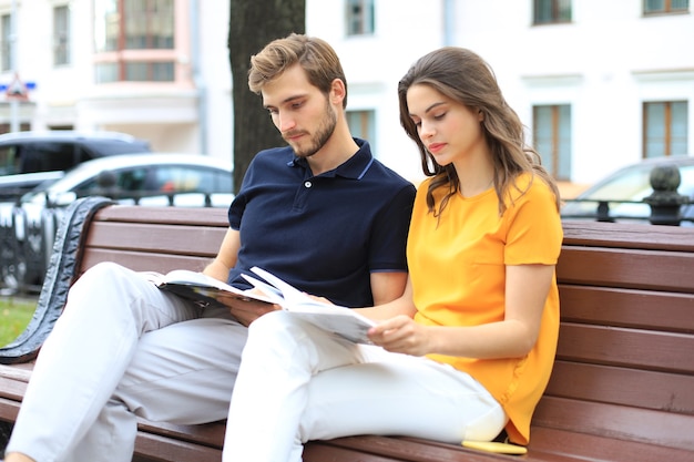 Casal jovem romântico com roupas de verão, sorrindo e lendo livros juntos enquanto está sentado no banco na rua da cidade.