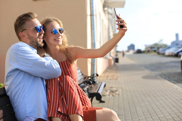 Casal jovem romântico com roupas de verão, sorrindo e tomando selfie enquanto está sentado no banco na rua da cidade.