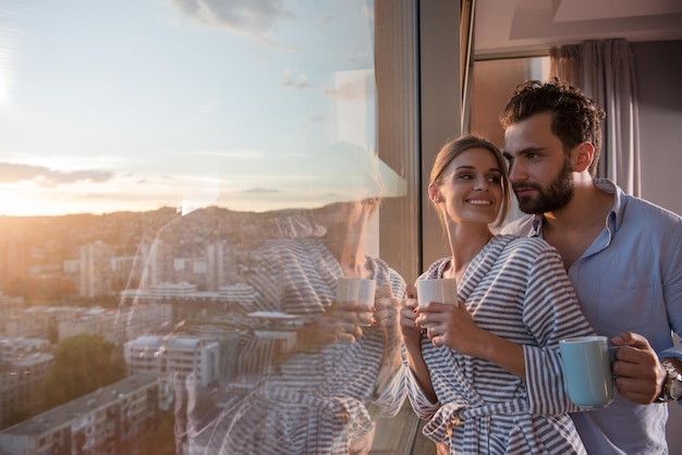 casal jovem romântico feliz desfrutando de café à noite e bela paisagem do pôr do sol da cidade em pé perto da janela