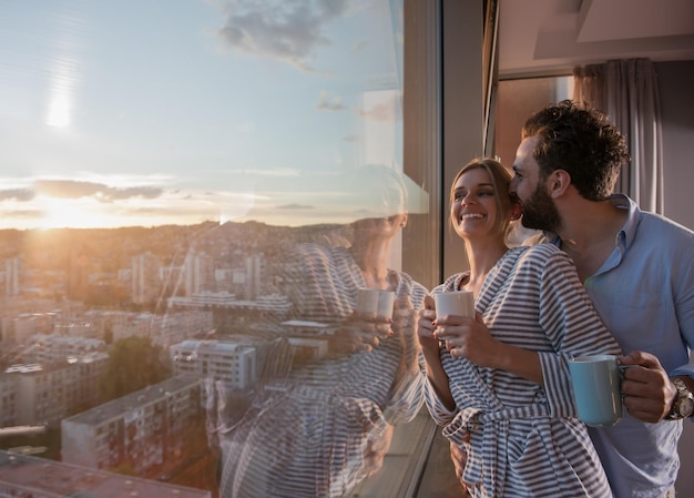 casal jovem romântico feliz desfrutando de café à noite e bela paisagem do pôr do sol da cidade em pé perto da janela