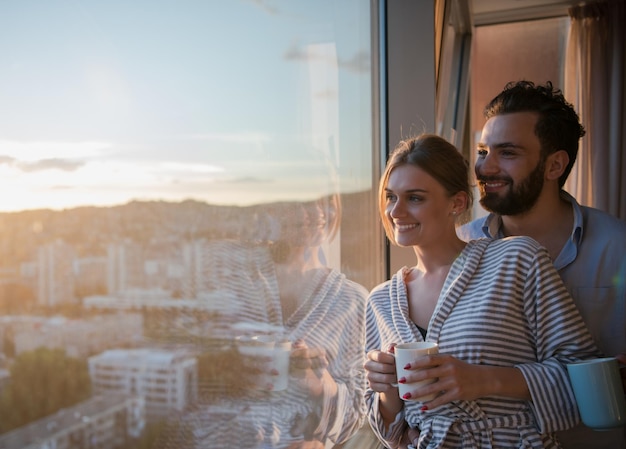 casal jovem romântico feliz desfrutando de café à noite e bela paisagem do pôr do sol da cidade em pé perto da janela