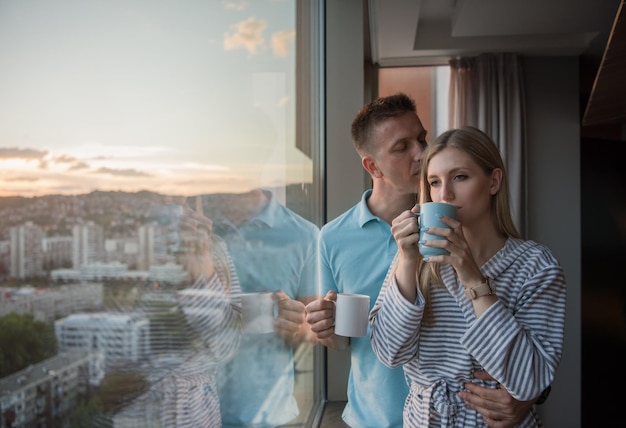 casal jovem romântico feliz desfrutando de café à noite e bela paisagem do pôr do sol da cidade em pé perto da janela