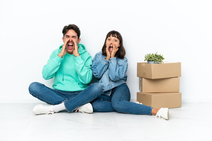 Foto casal jovem se mexendo enquanto pega uma caixa cheia de coisas sentado no chão, isolado no fundo branco, gritando e anunciando algo
