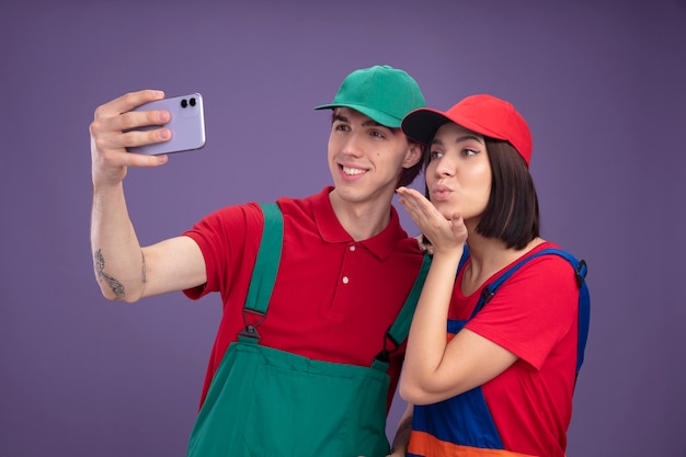 Casal jovem sorridente cara garota séria em uniforme de trabalhador da construção civil e boné tomando selfie juntos garota mandando golpe beijo isolado na parede roxa