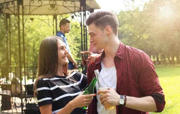 Casal jovem sorridente com cerveja e olhando um para o outro enquanto duas pessoas fazem churrasco no fundo.