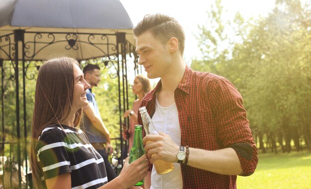 Casal jovem sorridente com cerveja e olhando um para o outro enquanto duas pessoas fazem churrasco no fundo.