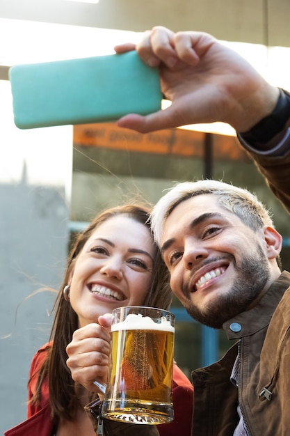 Foto casal jovem sorridente, desfrutando de uma cerveja juntos e tirando uma selfie com o celular. imagem vertical. conceito de tempo de lazer. conceito de tecnologia.