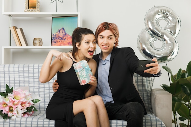 Casal jovem sorridente no dia da mulher feliz segurando e aponta no presente tirar uma selfie sentado no sofá da sala de estar