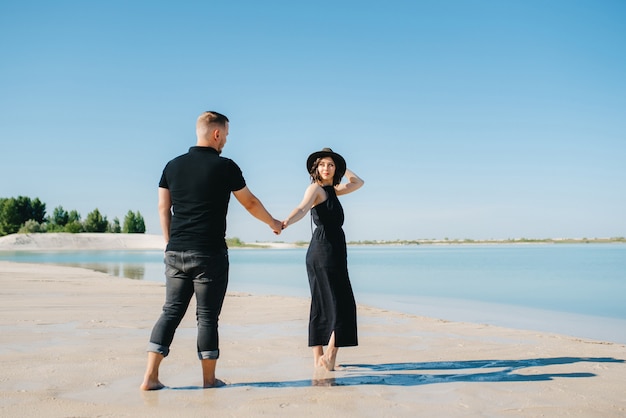 Casal jovem, um cara com uma garota em roupas pretas, está caminhando na areia branca à beira da água azul