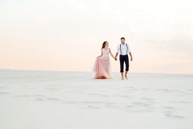 Casal jovem, um cara de calça preta e uma garota de vestido rosa, caminham pela areia branca do deserto