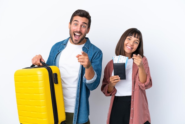 Casal jovem viajante segurando uma mala e passaporte isolado no fundo branco aponta o dedo para você com uma expressão confiante