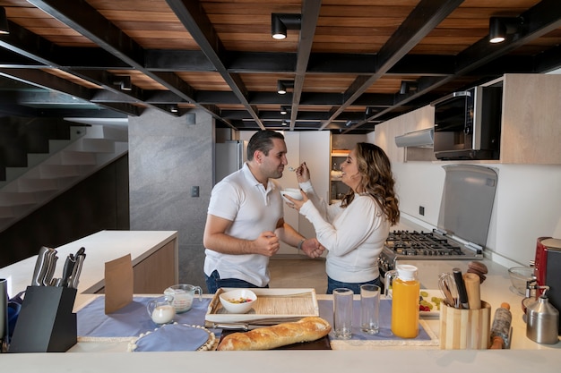 Foto casal latino feliz comendo iogurte com frutas enquanto prepara o café da manhã na cozinha.