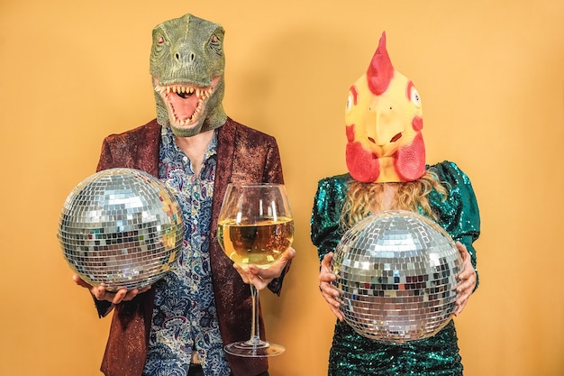 Casal louco se divertindo comemorando a festa de ano novo segurando bolas de discoteca e um copo de vinho - Foco em t-rex e máscara de frango