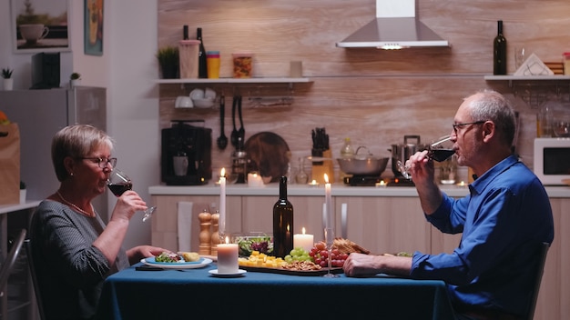 Casal maduro de idosos, bebendo uma taça de vinho durante o jantar romântico. idosos tilintando, sentados à mesa da cozinha, apreciando a refeição, comemorando seu aniversário na sala de jantar.